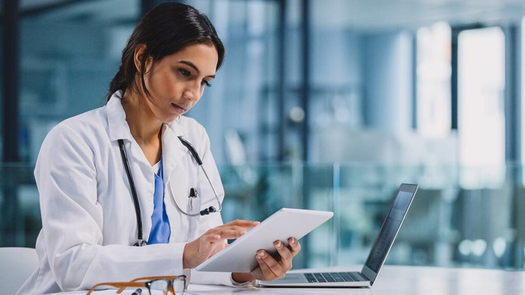 A doctor sitting next to her laptop uses her tablet.