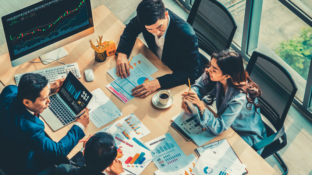 Business people sitting across from each other at a desk with a computer and clipboard with graphs and other information on it.